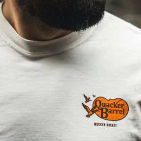 Close-up of a person wearing the Mucker Bucket Quacker Barrel Premium Tee, a relaxed fit garment-dyed cotton shirt with an orange and black flying ducks logo; part of their beard is visible above the light-colored tee.