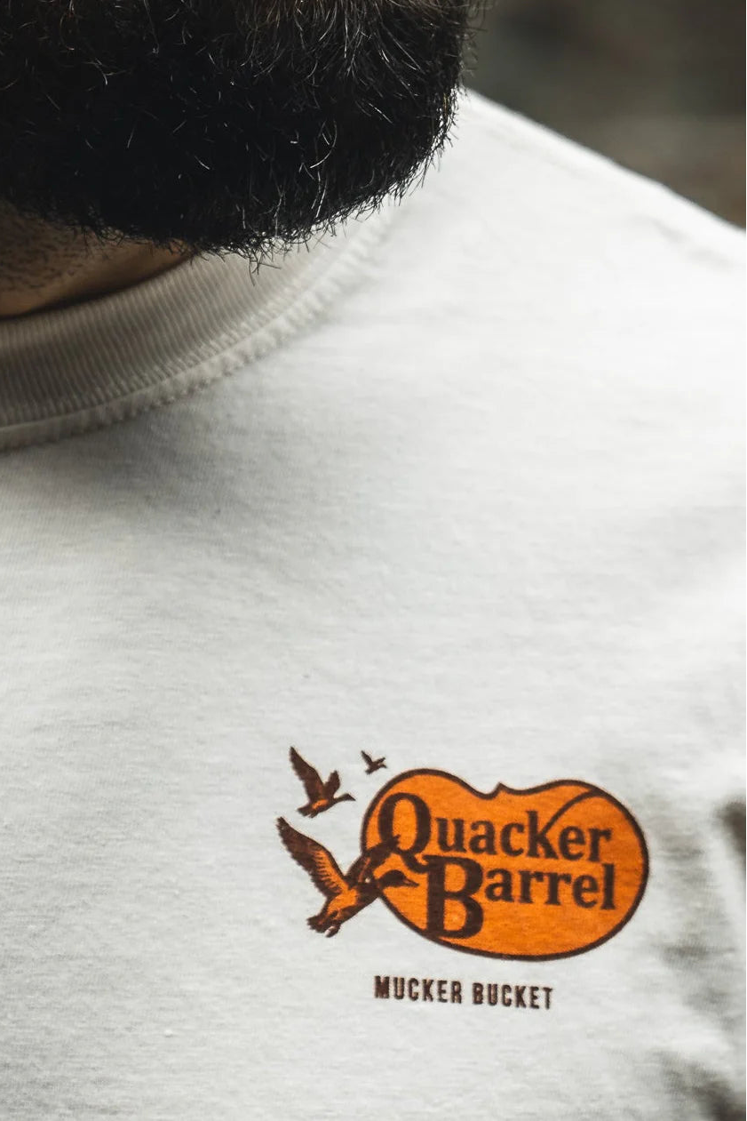 Close-up of a person wearing the Mucker Bucket Quacker Barrel Premium Tee, a relaxed fit garment-dyed cotton shirt with an orange and black flying ducks logo; part of their beard is visible above the light-colored tee.