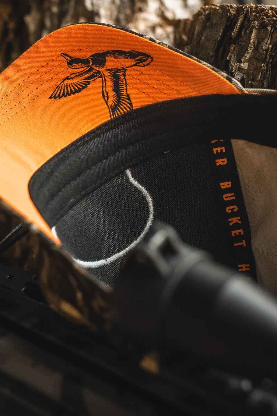 The Mucker Bucket Mallard Life Deadwood Camo Bucket features a bird illustration and orange brim, shown up close on hunting gear and wood—a perfect nod to mallard life.