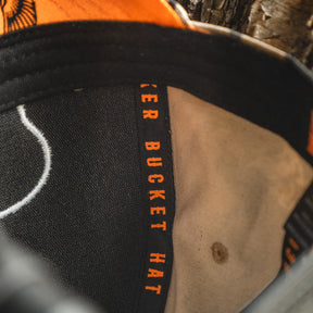 Close-up of the inside of the My Store Mallard Life Deadwood Camo Bucket hat, featuring orange "BUCKET HAT" text on the inner band and a worn, textured camo fabric inspired by mallard life.