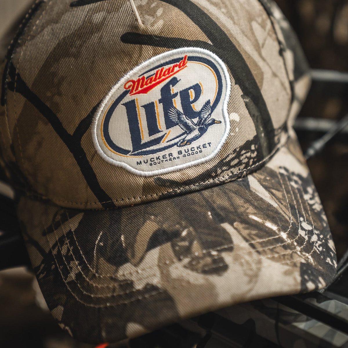 The Mallard Life Deadwood Camo Bucket by My Store is a camo snapback hat with a patch that reads "Mallard Life Mucker Bucket Southern Goods," including a flying duck graphic and brown-tan accents.