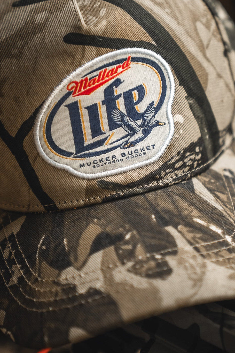 A close-up of the Mallard Life Deadwood Camo Bucket by Mucker Bucket, featuring a patch with a duck graphic and text, resting on a dark surface.