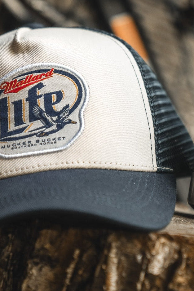 The Mucker Bucket Mallard Life Trucker Bucket is a beige and black mallard hat with a Miller Lite patch, bird illustration, and mesh back snapback, shown on a rough wooden surface.