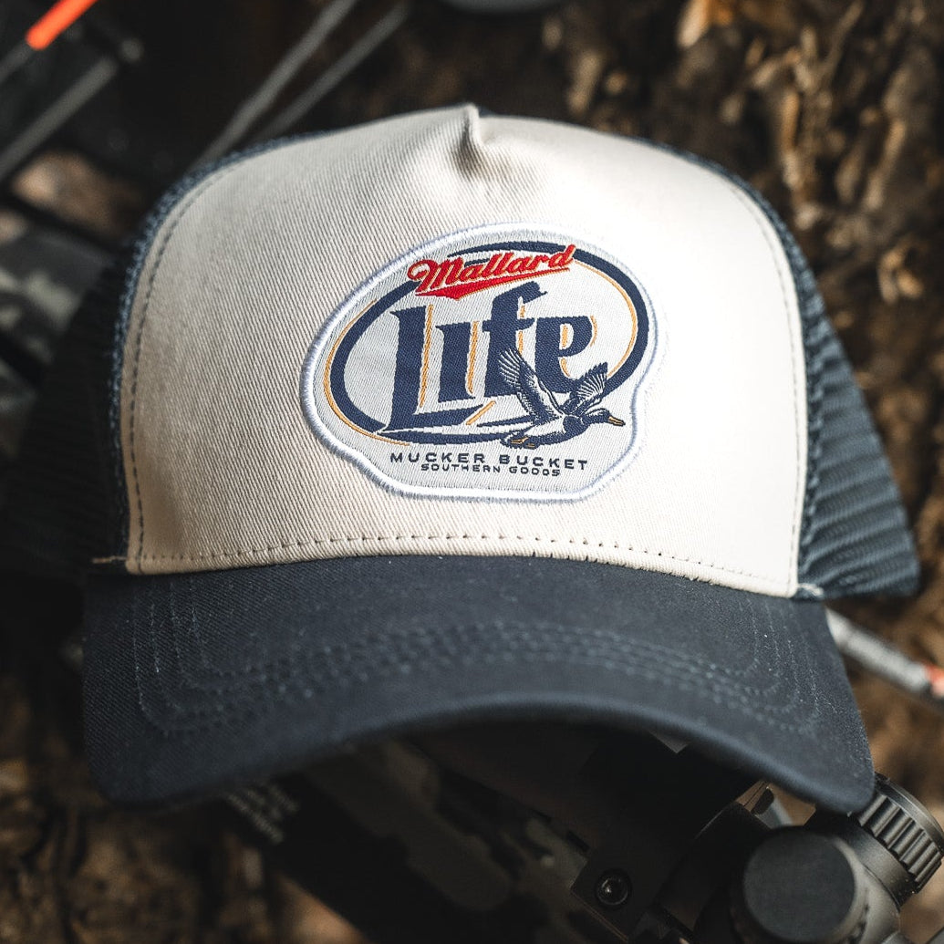 The Mallard Life Trucker Bucket from My Store, a navy and white snapback hat with patch embroidery of two flying ducks and "Mallard Life," sits on dark equipment outdoors against a natural backdrop.
