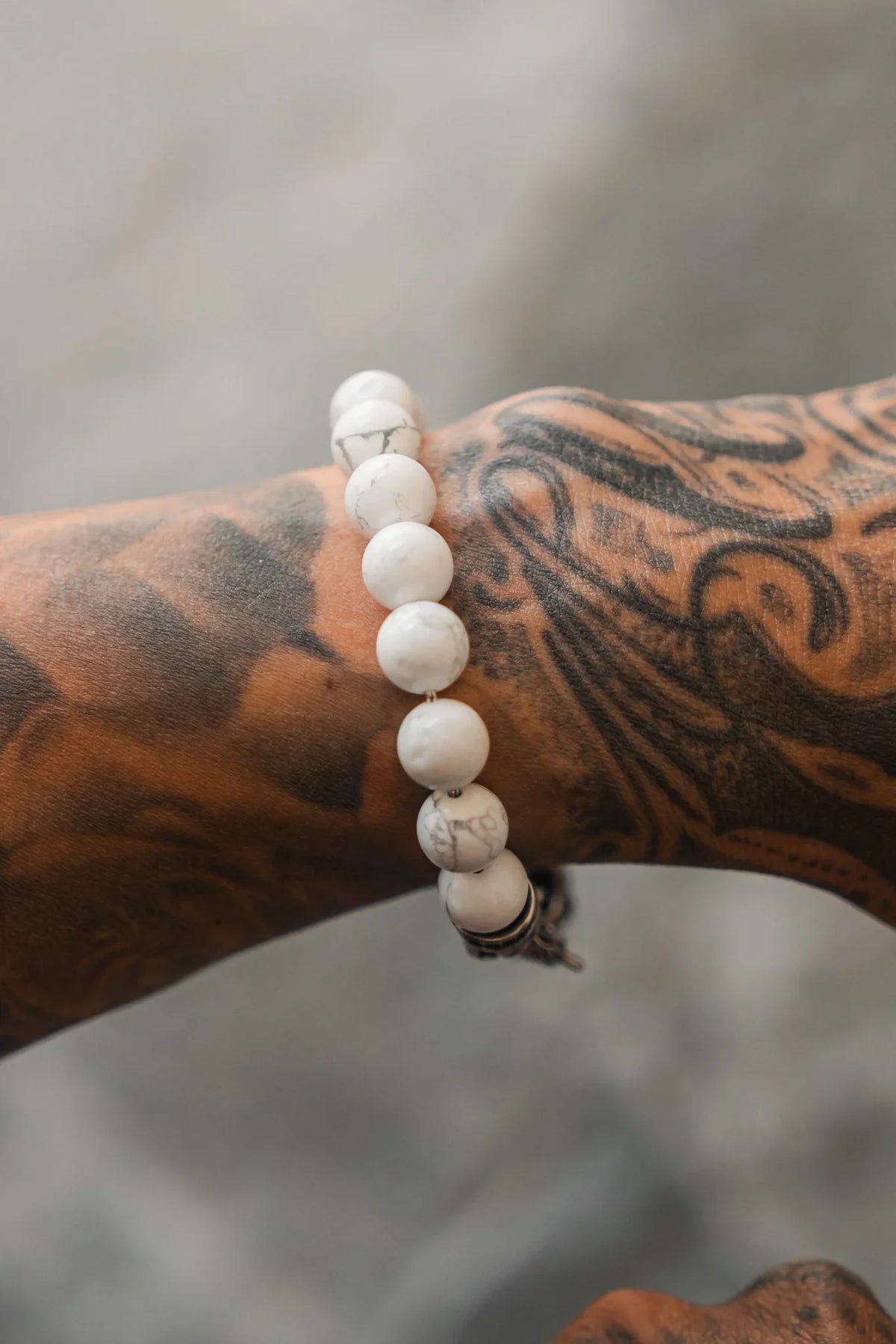 A close-up of a wrist with a black tattoo, wearing the Relentless Betrayal Storm Cloud Gemstone Bracelet—white beads with marbled patterns, semi-precious gemstones, and a stainless steel clasp—set against a blurred background.