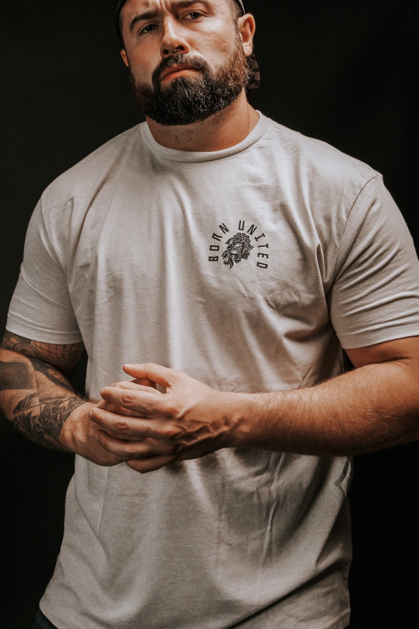 A bearded, tattooed man stands against a dark background, wearing a light gray Beware T-shirt by Born United Private Label and a backward baseball cap. With clasped hands and a focused look, he exudes mystery, as if discerning hidden truths beyond false facades.