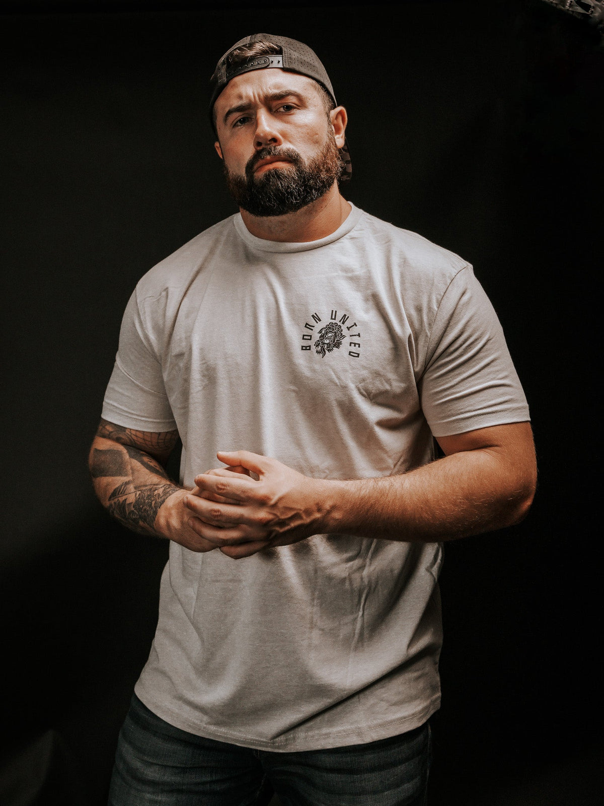 A bearded, tattooed man stands against a dark background, wearing a light gray Beware T-shirt by Born United Private Label and a backward baseball cap. With clasped hands and a focused look, he exudes mystery, as if discerning hidden truths beyond false facades.