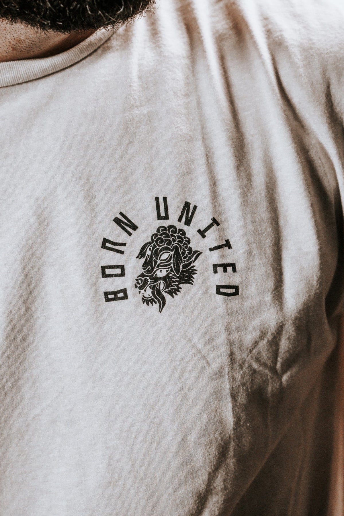 Close-up of someone wearing a white Born United Private Label Beware T-shirt, featuring BORN UNITED and a lion graphic on the chest against a dark background. The bold design stands out, warding off false prophets in sheeps clothing.