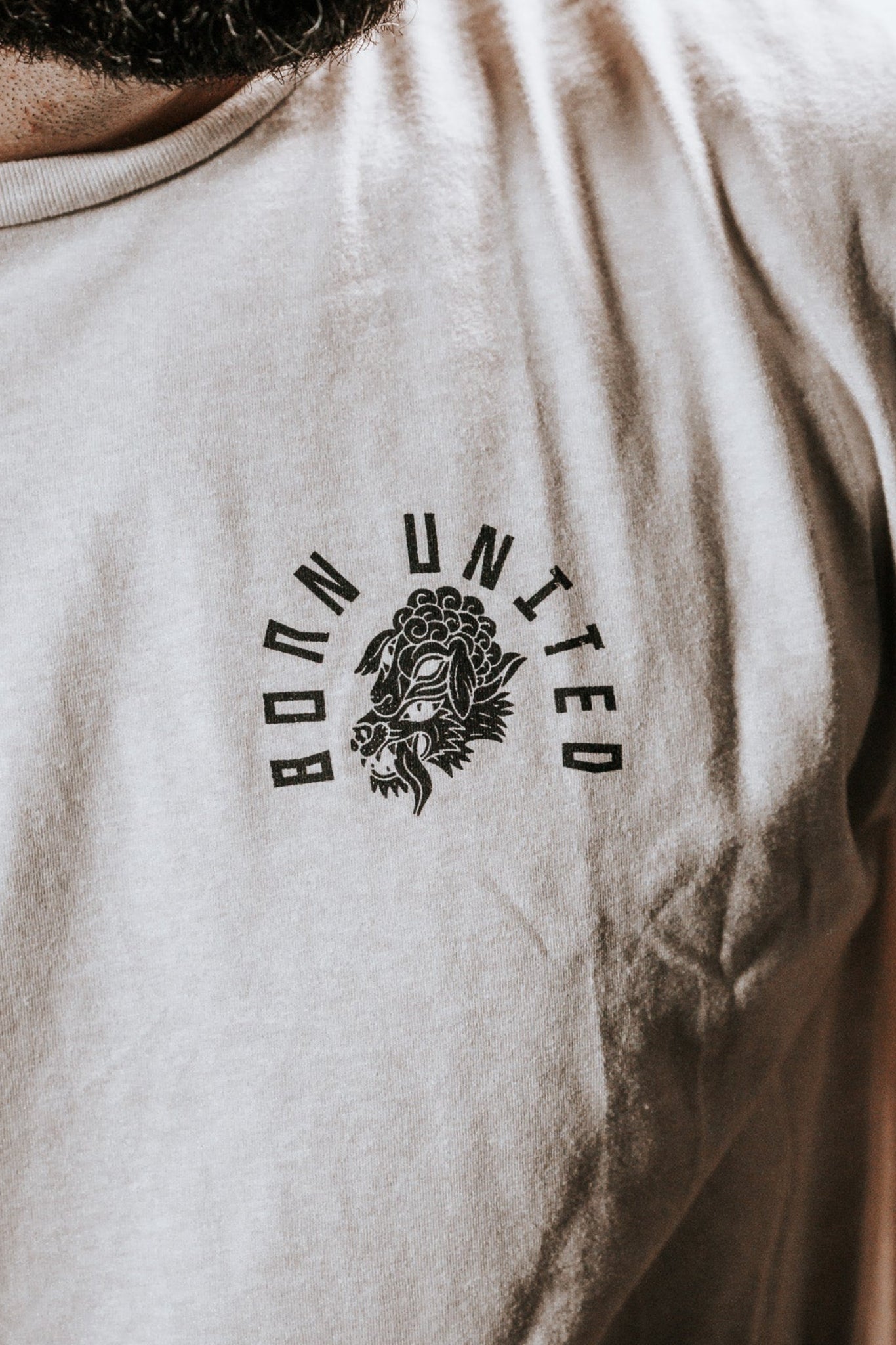 Close-up of someone wearing a white Born United Private Label Beware T-shirt, featuring BORN UNITED and a lion graphic on the chest against a dark background. The bold design stands out, warding off false prophets in sheeps clothing.