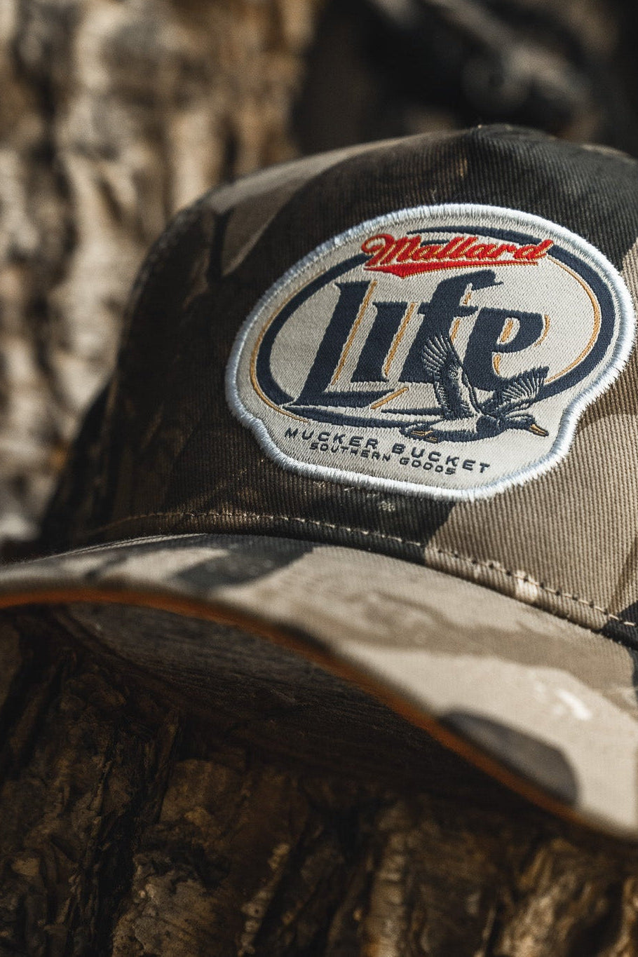 The Mucker Bucket Mallard Life Deadwood Camo Bucket, featuring a flying duck patch, rests on a tree trunk outdoors.