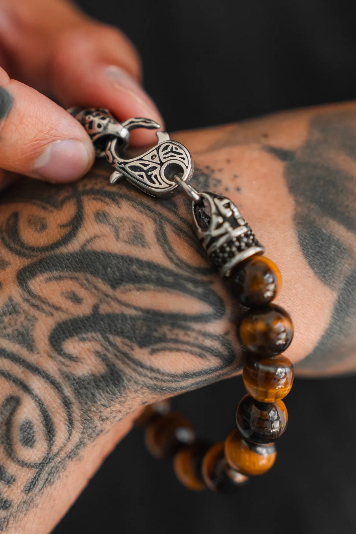 A tattooed hand fastens the Relentless Betrayal Tigers Eye Gemstone Bracelet, featuring brown beads and an ornate silver clasp, emphasizing its striking details and protective metaphysical properties.
