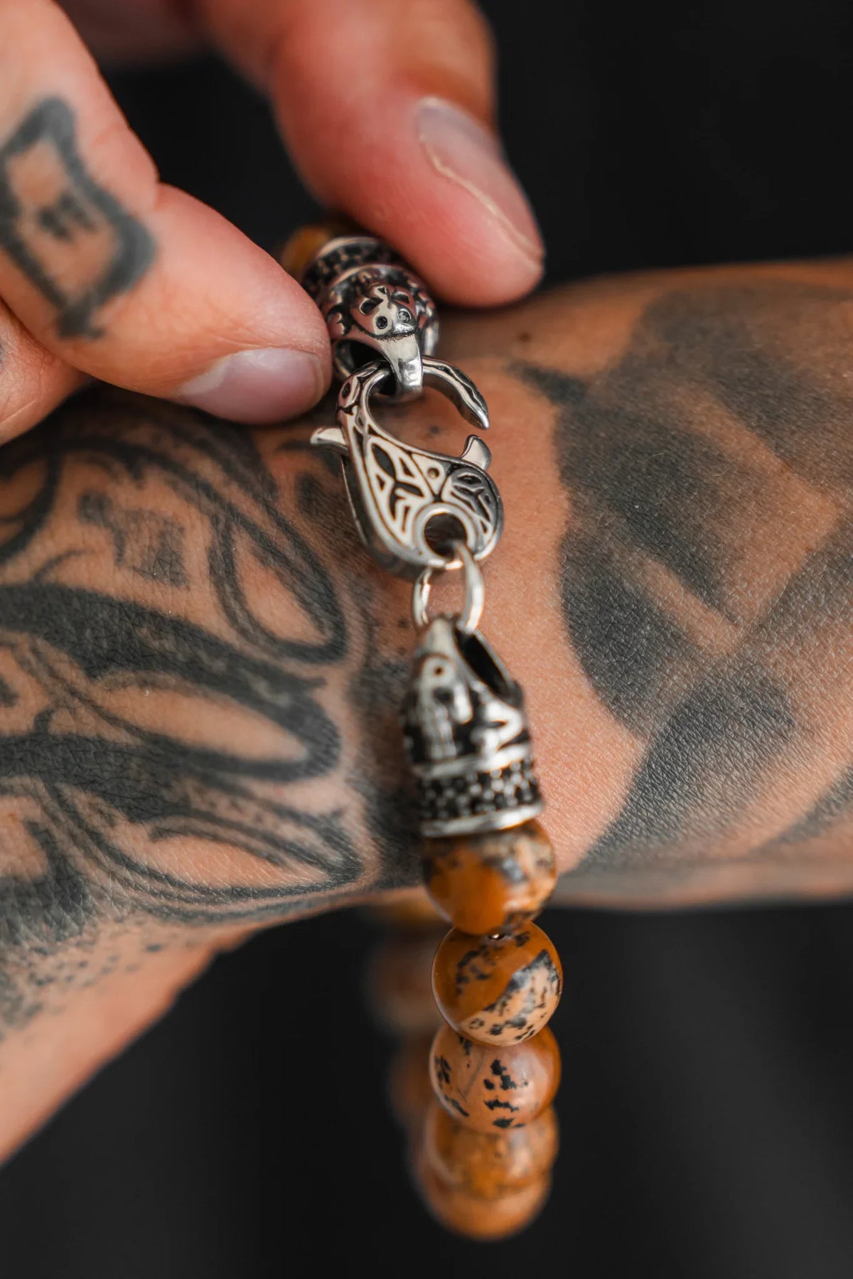 A tattooed hand fastens a detailed stainless steel clasp on the Relentless Betrayal Desert Storm Gemstone Bracelet, which features brown marbled semi-precious stones and ornate silver accents, against a tattooed wrist.