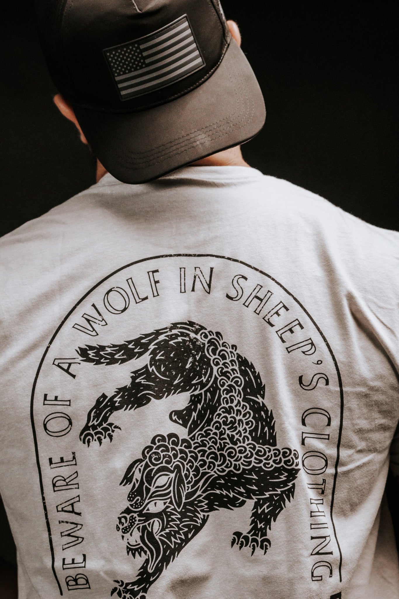 A person wearing a black cap with an American flag and a gray Beware shirt by Born United Private Label, featuring a wolf illustration, stands facing away. The shirt warns against false prophets with the phrase Beware of a Wolf in Sheeps Clothing.