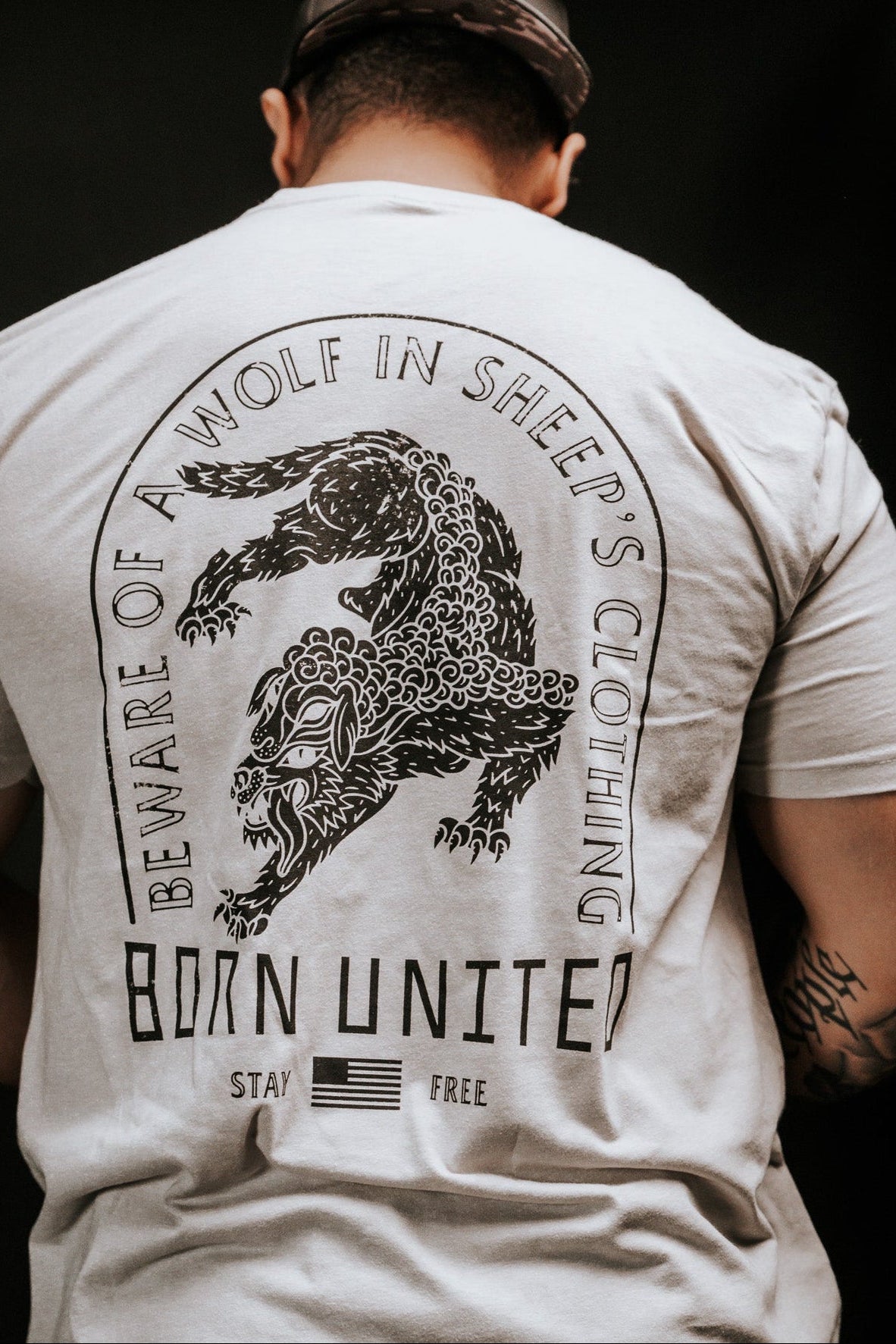 A person wears a light-colored t-shirt with a snarling wolf illustration and the words Beware of ravening wolves in sheeps clothing, plus Born United, Stay Free, and a small American flag. The brand is Born United Private Label, and the product name is Beware.