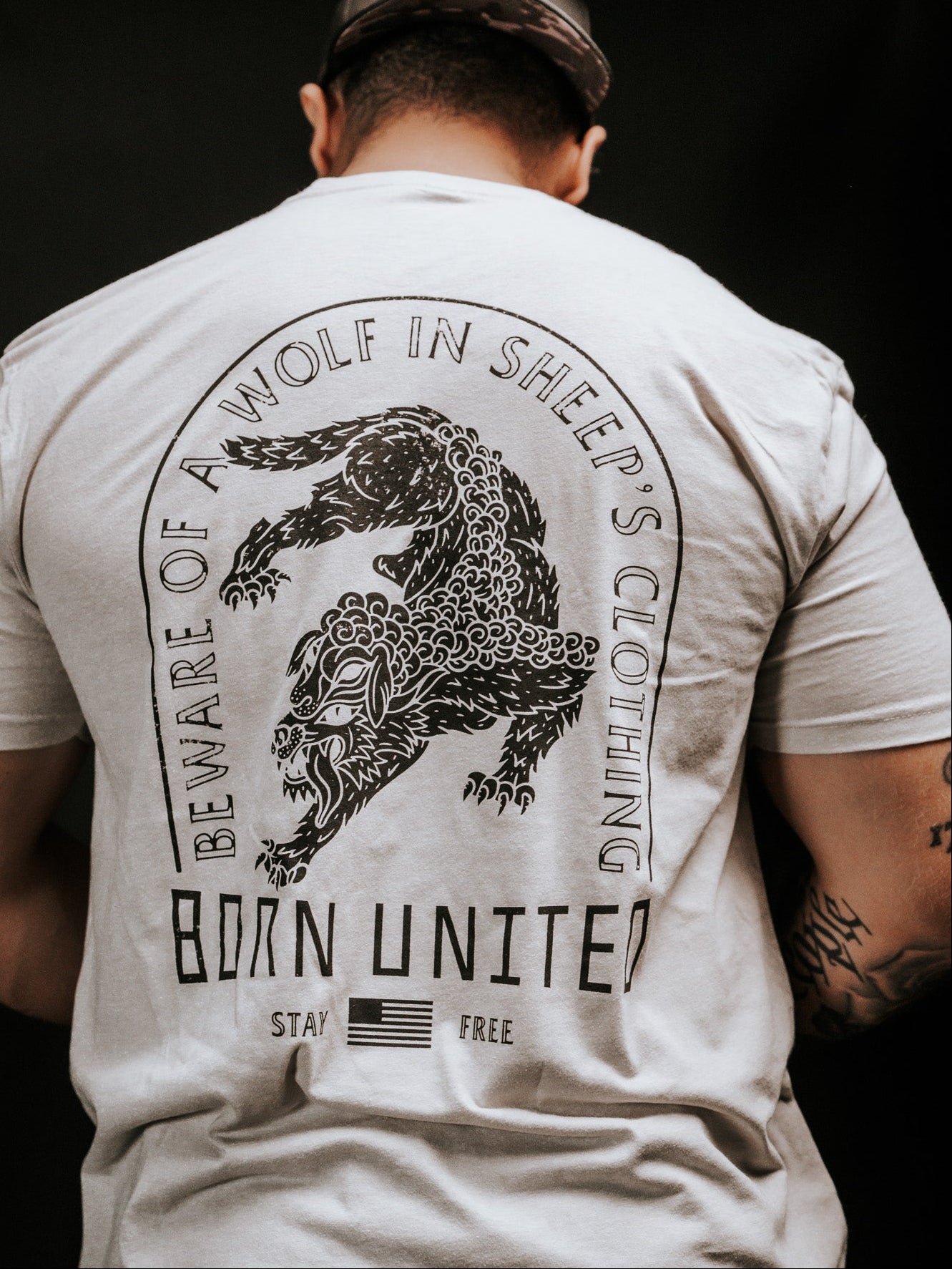 A person wears a light-colored t-shirt with a snarling wolf illustration and the words Beware of ravening wolves in sheeps clothing, plus Born United, Stay Free, and a small American flag. The brand is Born United Private Label, and the product name is Beware.