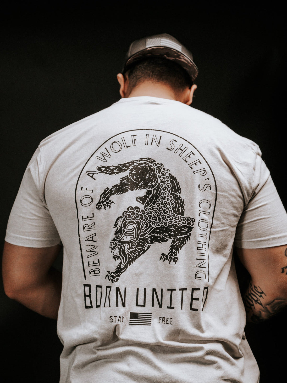 A person wears a light-colored t-shirt with a snarling wolf illustration and the words Beware of ravening wolves in sheeps clothing, plus Born United, Stay Free, and a small American flag. The brand is Born United Private Label, and the product name is Beware.