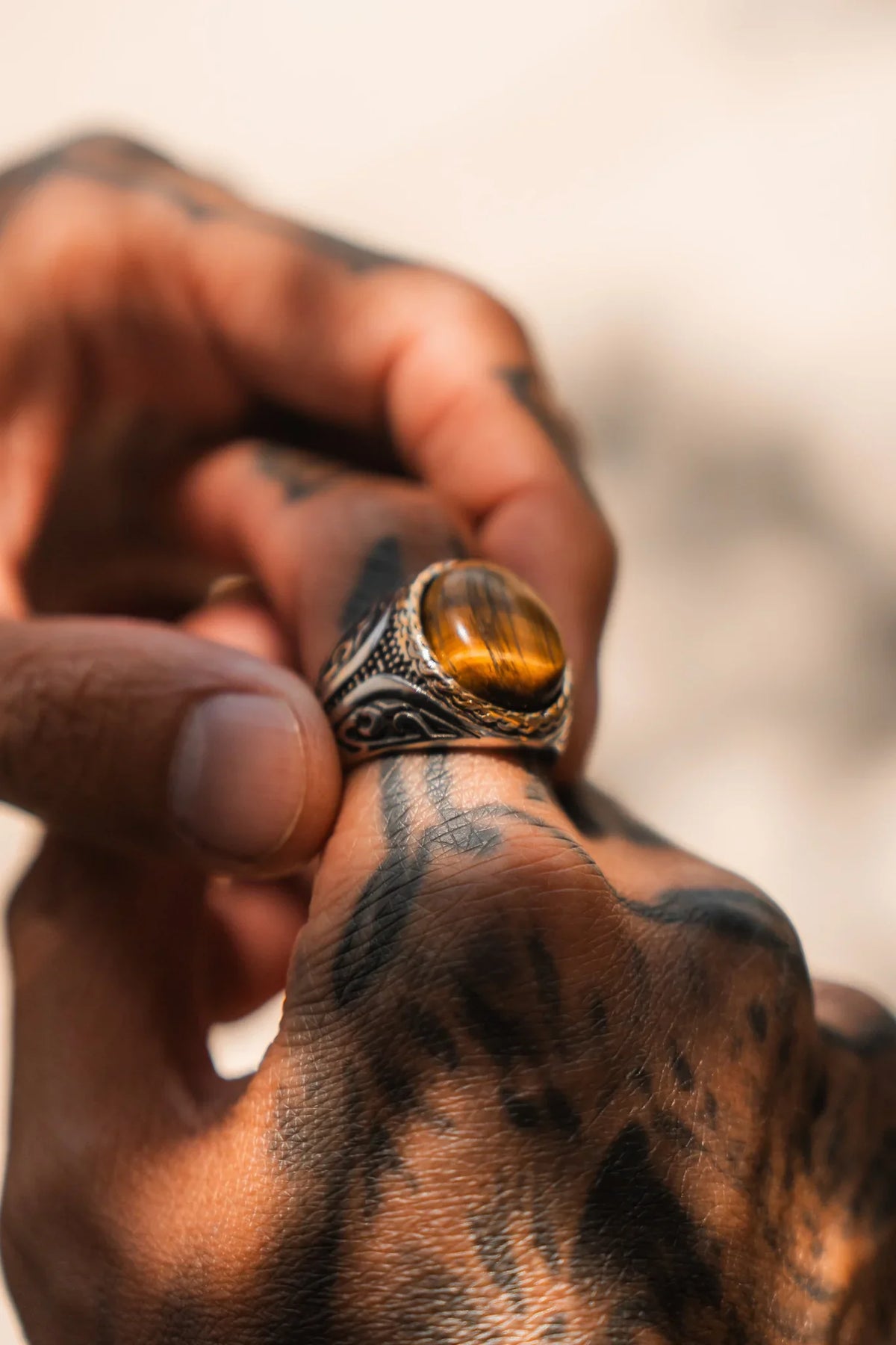 A tattooed hand holds the Relentless Betrayal Imperial Tiger’s Eye Ring—a handcrafted stainless steel piece featuring an oval tiger’s eye gemstone—set against a softly blurred light background.