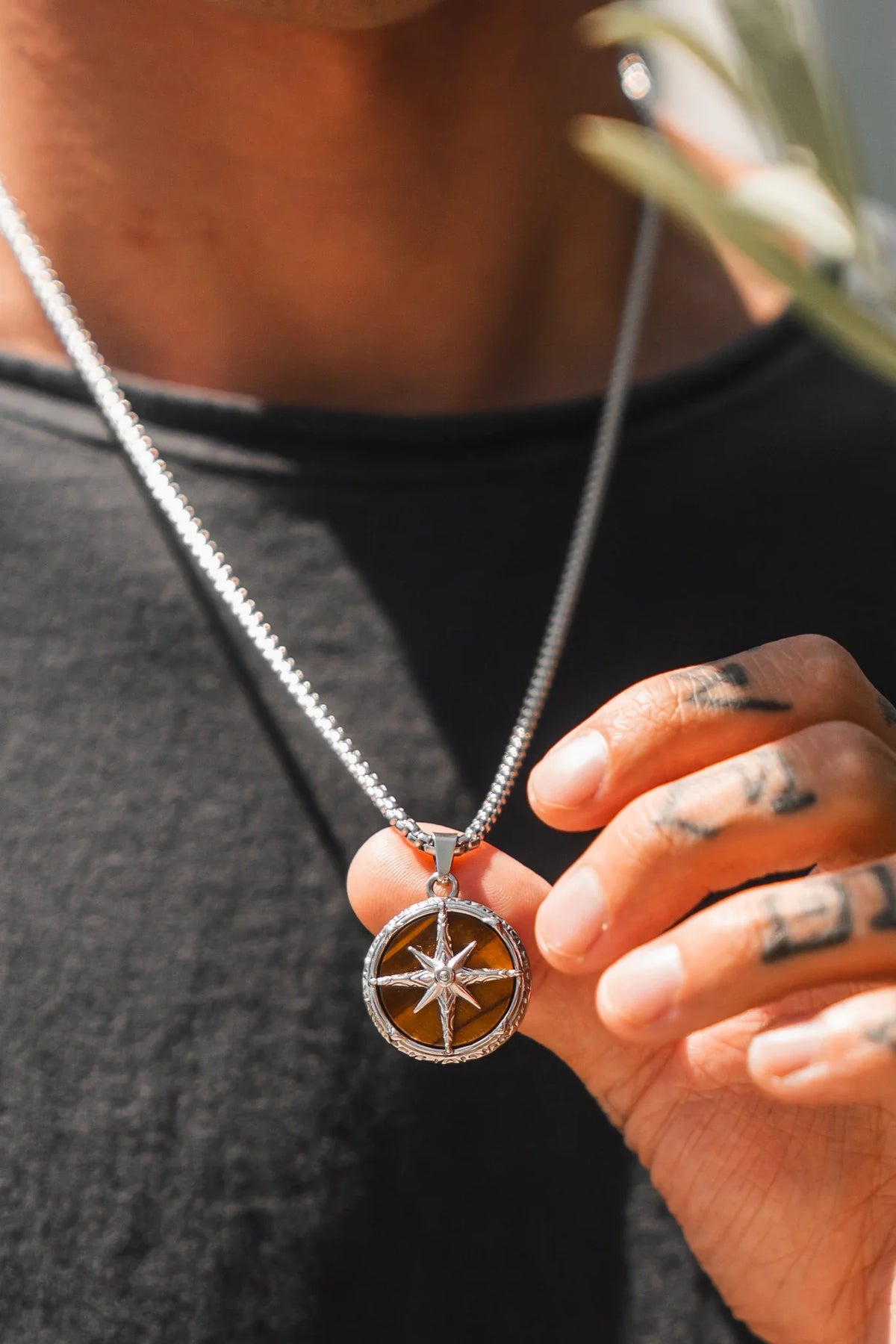 A person in a black shirt displays the Relentless Betrayal Guide Of The Tiger Necklace, showcasing the handcrafted silver pendant with a Tigers Eye stone and star design as sunlight highlights its brown background. Their tattooed hand holds the jewelry.