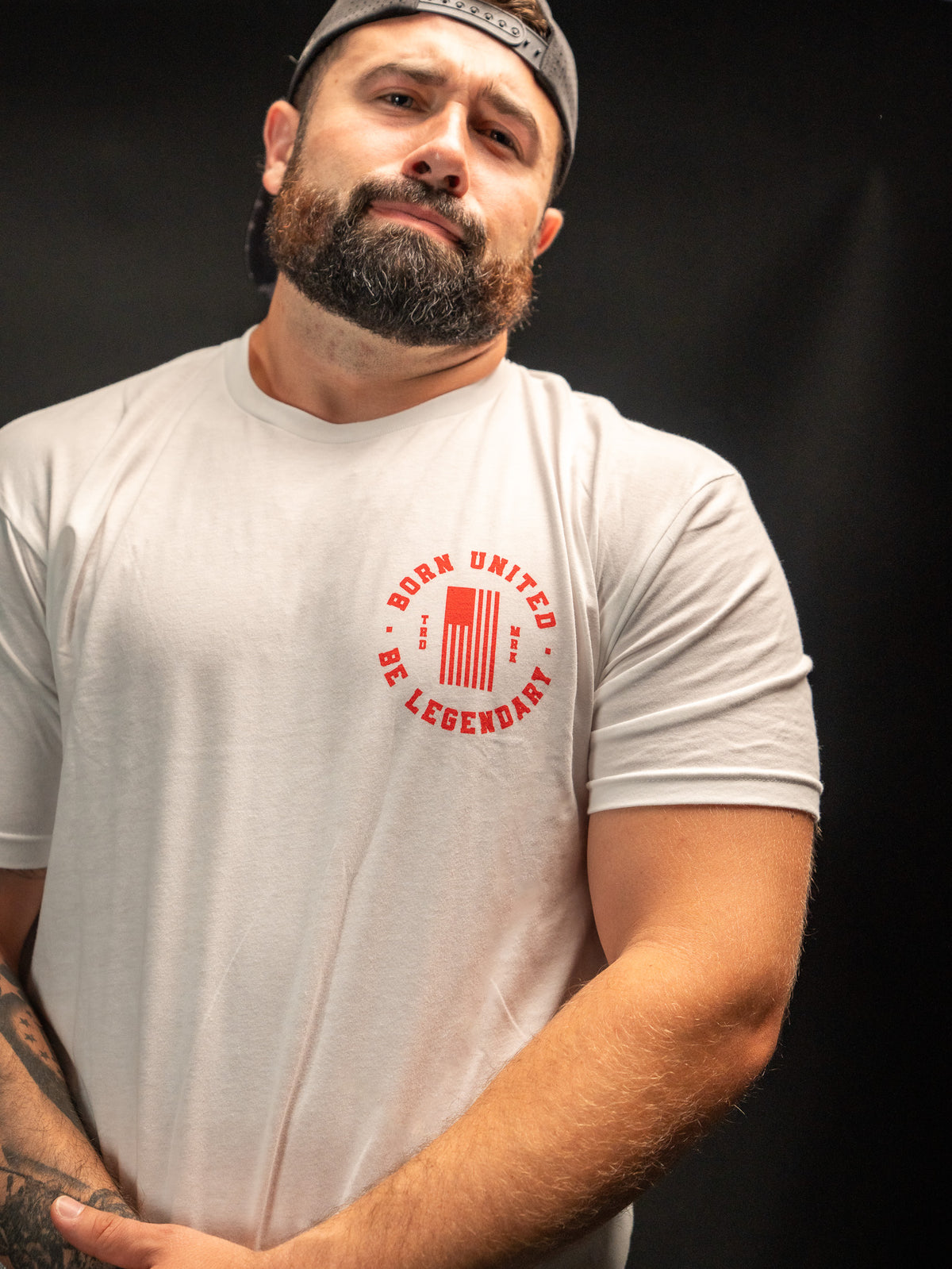 A bearded, capped man wearing a Born United Private Label t-shirt with Born United, Be Legendary in red text poses with a raised eyebrow against a black background, channeling the timeless charm of Legends - Dukes of Hazzard.