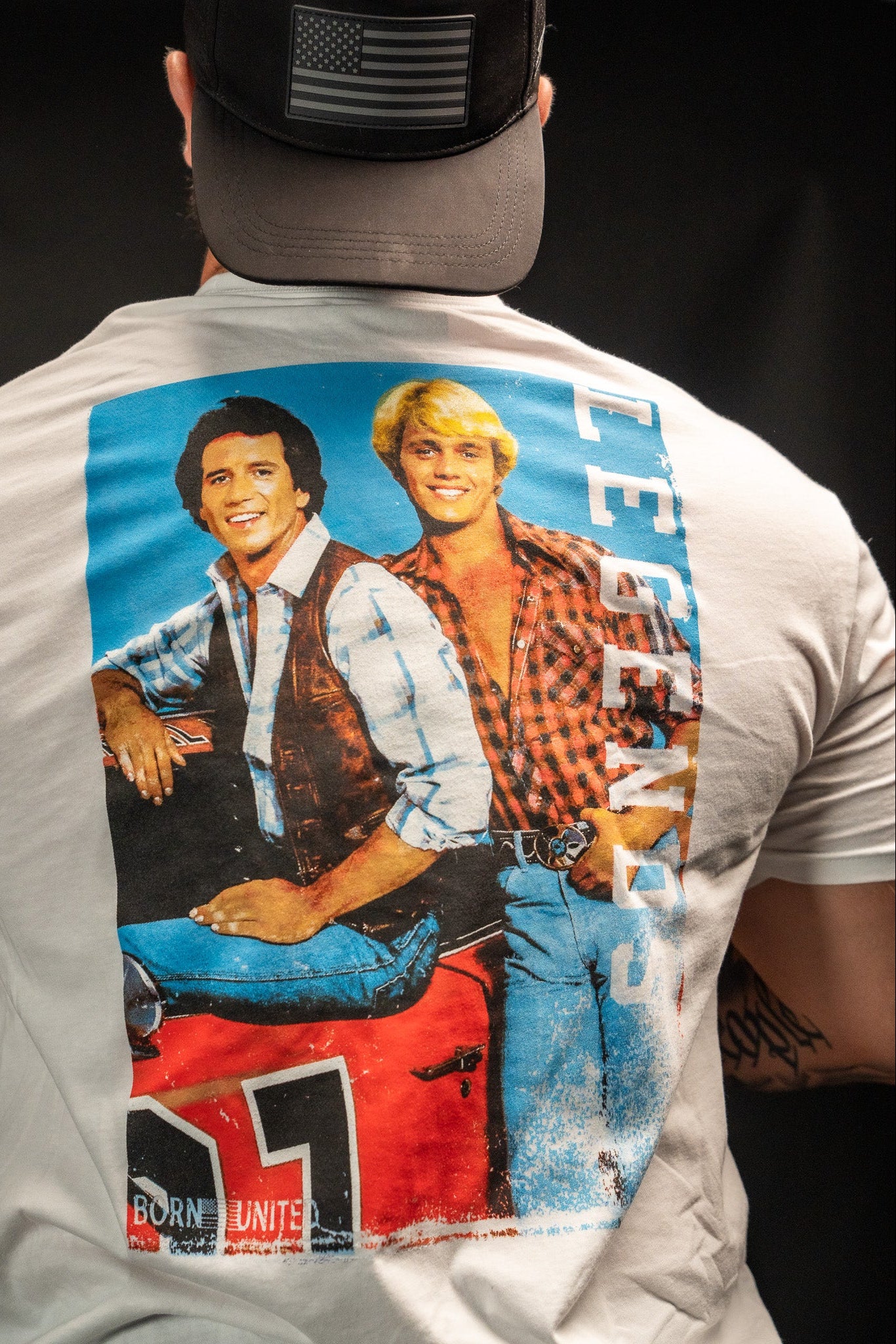 The image shows someone wearing the Born United Private Labels Legends - Dukes of Hazzard white T-shirt, featuring two smiling good old boys and a red car, with LEGENDS printed vertically. They also have a black cap with an American flag, embodying that timeless spirit of never meaning no harm.