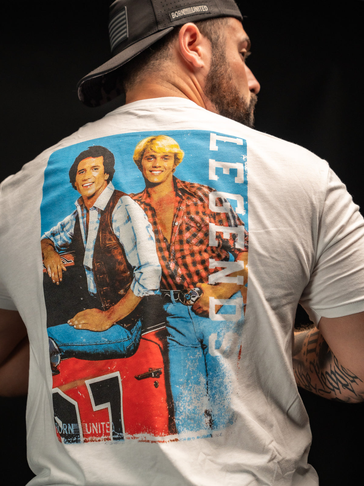 A man sports a white T-shirt from Born United Private Label featuring a retro Legends - Dukes of Hazzard graphic with two people and the word LEGENDS in blue. He embodies the classic good old boys vibe, slightly turned away and wearing a slick black cap.