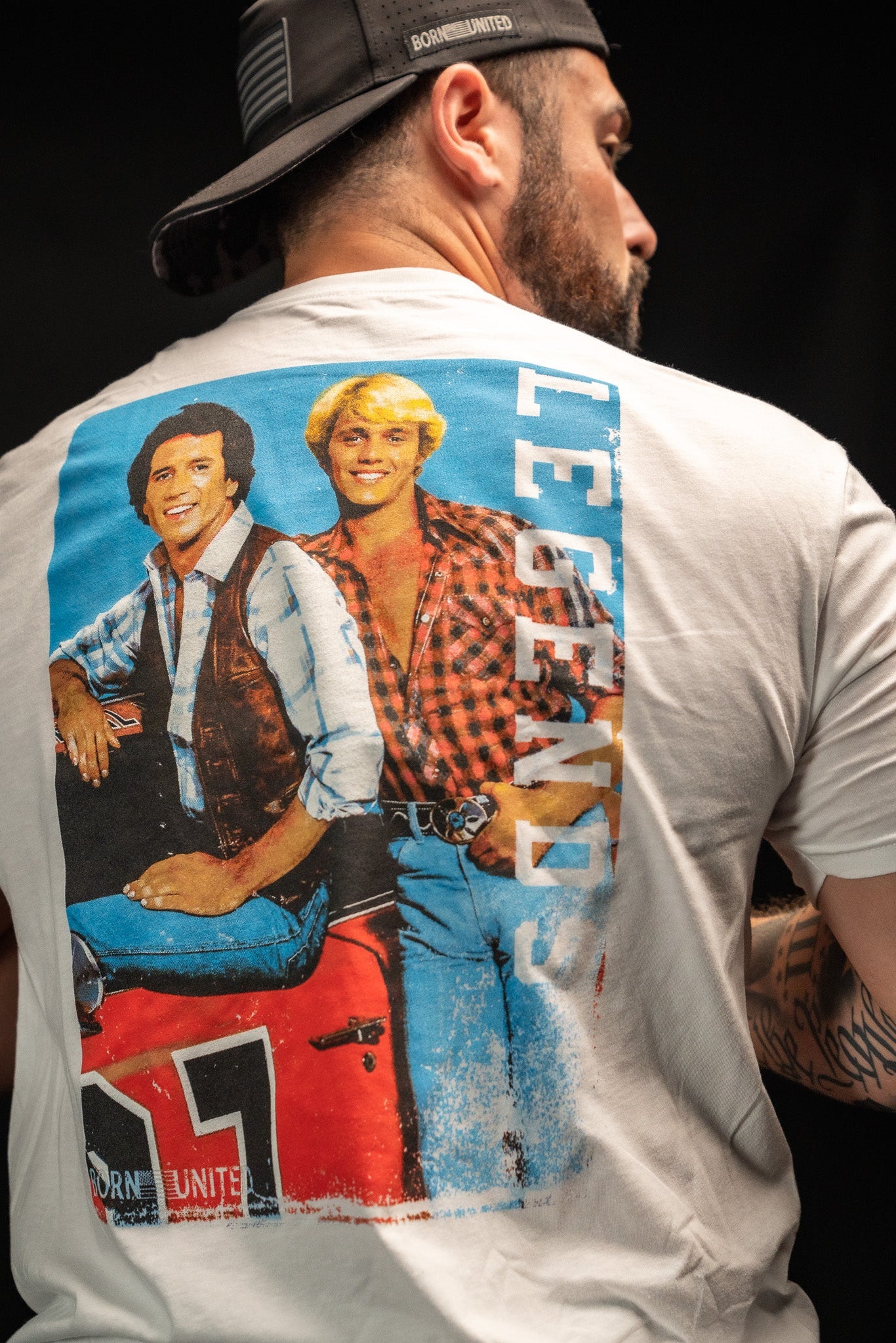 A man sports a white T-shirt from Born United Private Label featuring a retro Legends - Dukes of Hazzard graphic with two people and the word LEGENDS in blue. He embodies the classic good old boys vibe, slightly turned away and wearing a slick black cap.