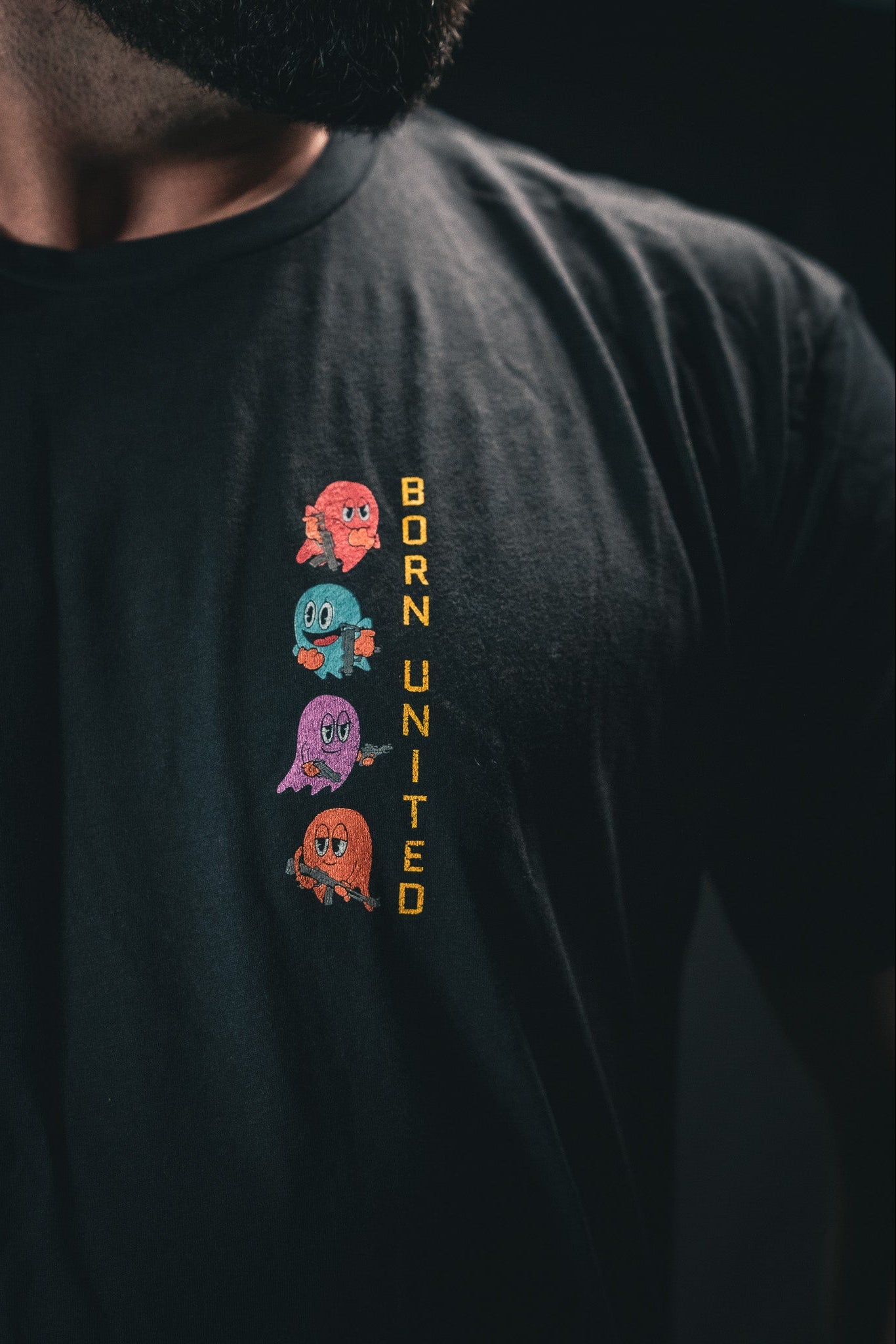 A person is wearing a Tac-Man (Warehouse) T-shirt from Born United Private Label. It features four vibrant cartoon ghosts stacked vertically on a dark backdrop, with BORN UNITED in yellow beside them.