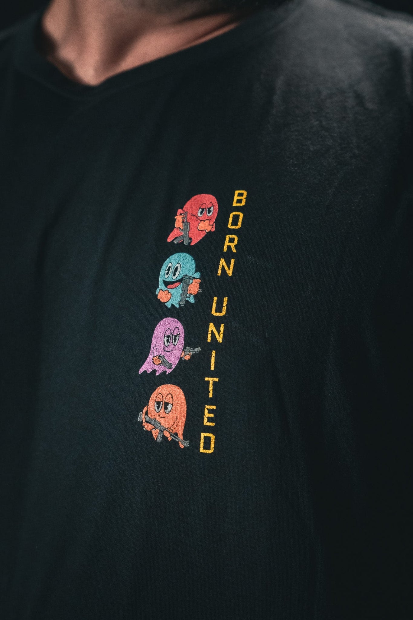 Someone is wearing a Tac-Man (Warehouse) T-shirt from Born United Private Label, featuring colorful ghost-like characters stacked vertically on the left side, with BORN UNITED written alongside.