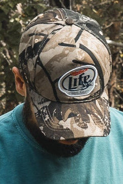 A person wearing a Mallard Life Deadwood Camo Bucket by Mucker Bucket, looking down in a teal shirt, surrounded by trees and foliage.