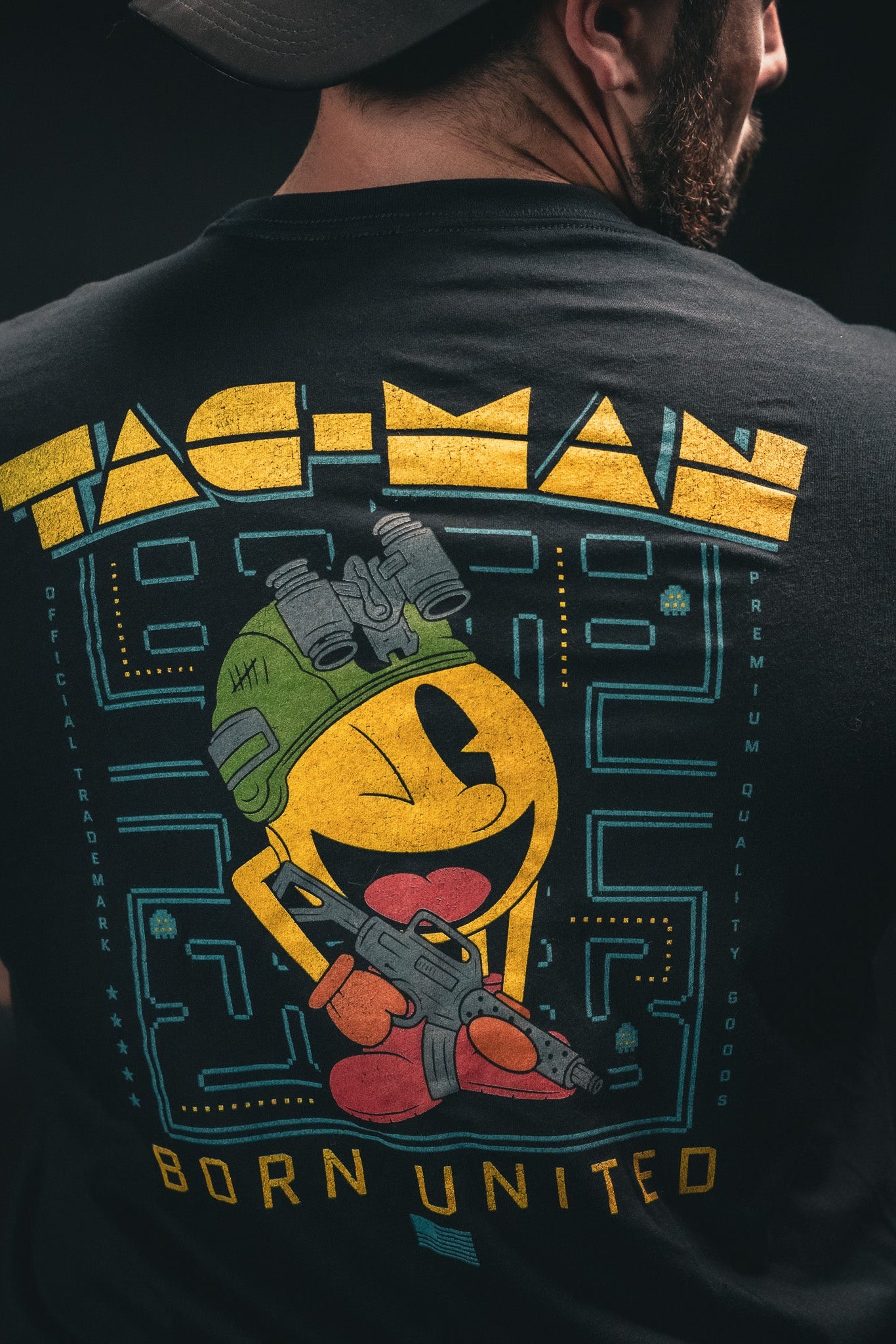 A man wears a black T-shirt featuring a yellow, round character in a helmet and boots, holding a gun. Tac-Man is above and Born United Private Label below the character, all in retro video game style.
