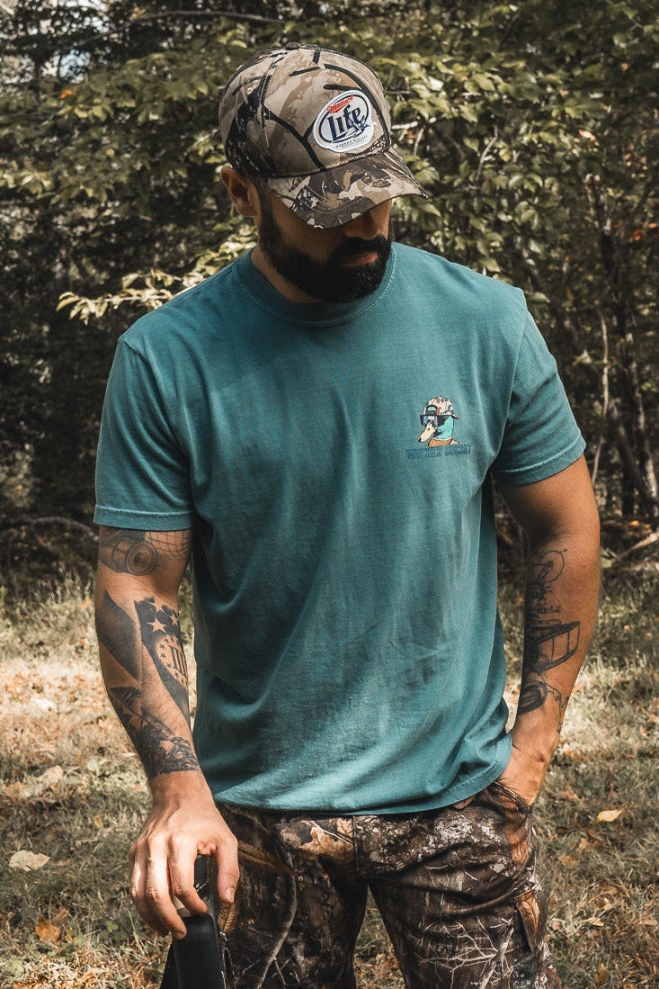 A bearded, tattooed man in a teal t-shirt stands outdoors in a wooded area wearing the Mucker Bucket Mallard Life Deadwood Camo Bucket hat and pants, looking down while holding a black object in one hand.