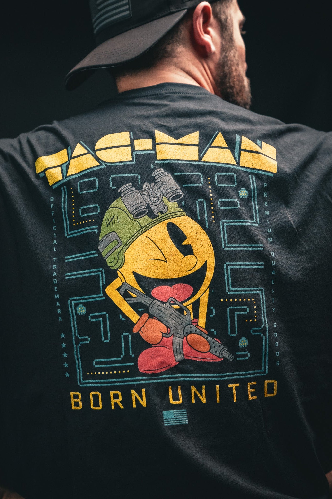 A person wears a black Tac-Man (Warehouse) T-shirt by Born United Private Label, featuring a yellow character with an army helmet and rifle against a maze. The text PAC-MAN is above and BORN UNITED below; theyre shown from the back.