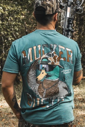 A man outdoors holds a hunting bow while wearing the Mucker Bucket Cool Duck Premium Tee, a garment-dyed, relaxed fit teal shirt featuring a duck in sunglasses and camo hat with "MUCKER DUCKER" on the back. Trees are behind him.