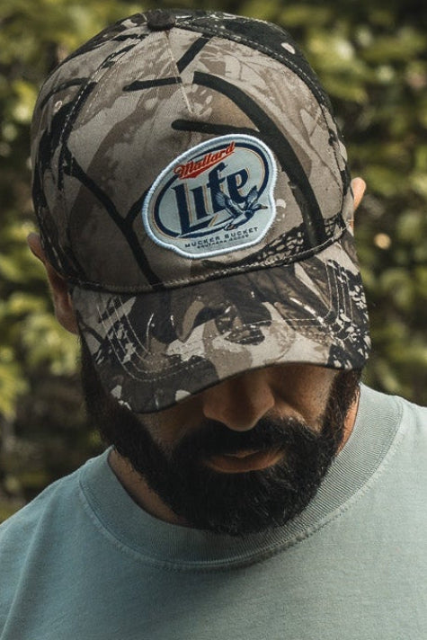 A bearded man looks down outdoors in greenery, wearing a light blue shirt and a Mucker Bucket Mallard Life Deadwood Camo Bucket hat.