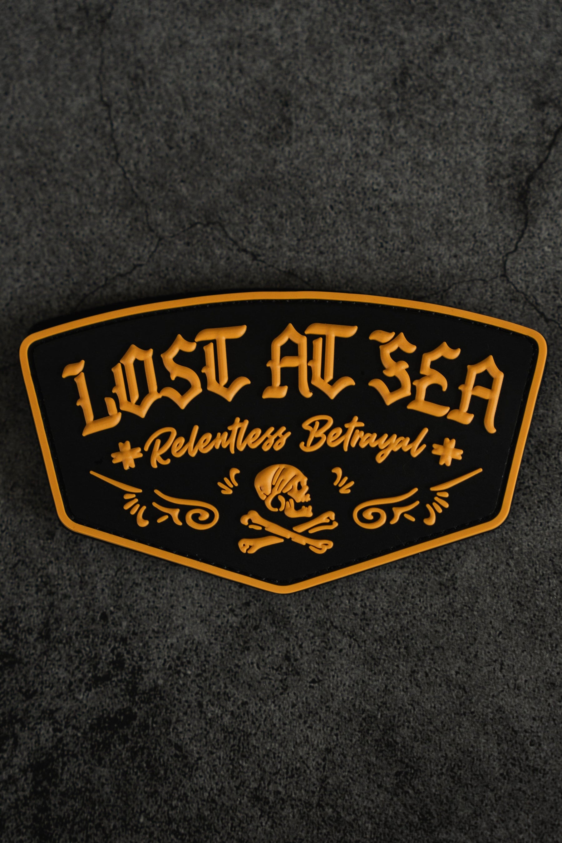 A black and gold Lost At Sea PVC Patch by Relentless Betrayal, featuring a bold skull and crossbones design on a textured dark background—the perfect accessory for your Lost at Sea hoodie or survival hoodie.