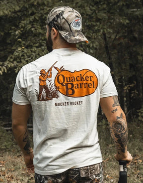 A tattooed man in a camo hat and pants stands outdoors, wearing the Quacker Barrel Premium Tee by Mucker Bucket—crafted from garment dyed cotton, it features an orange hunter graphic and "MUCKER BUCKET" beneath the logo.