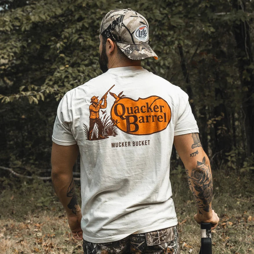 A tattooed man in a camo hat and pants stands outdoors, wearing the Quacker Barrel Premium Tee by Mucker Bucket—crafted from garment dyed cotton, it features an orange hunter graphic and "MUCKER BUCKET" beneath the logo.