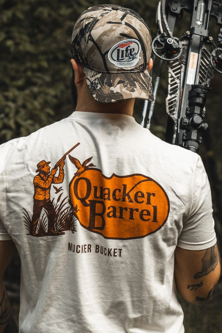 Wearing a Mucker Bucket Mallard Life Deadwood Camo Bucket hat and a Quacker Barrel T-shirt with a hunter and duck graphic, someone stands outdoors with a crossbow over their shoulder, surrounded by trees.