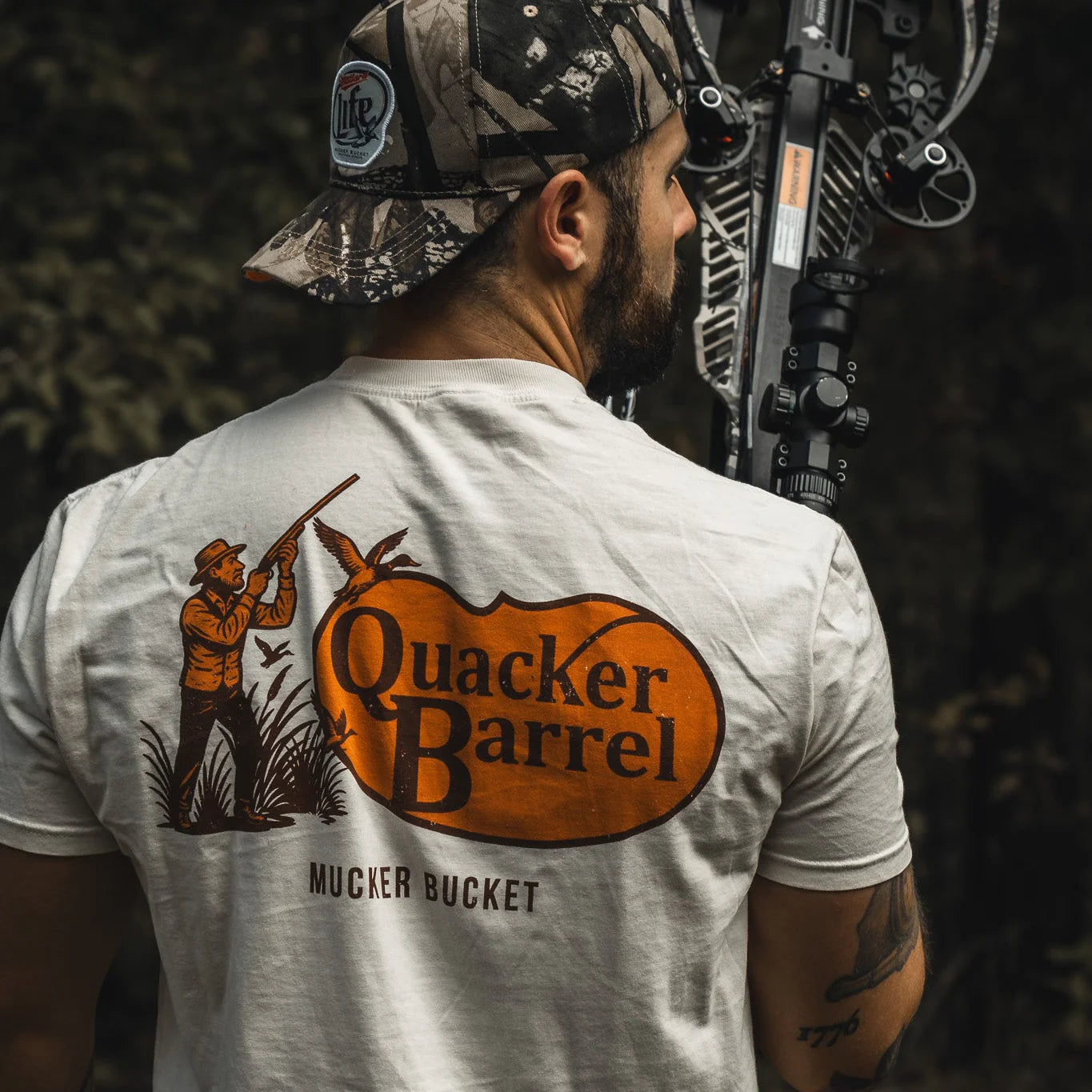 A man in a camouflage cap holds a compound bow outdoors, wearing the Mucker Bucket Quacker Barrel Premium Tee—garment-dyed cotton with a hunter, ducks, and “Quacker Barrel MUCKER BUCKET” graphic.