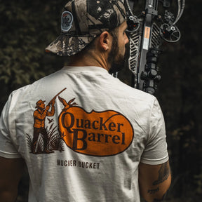 A man in a camouflage cap holds a compound bow outdoors, wearing the Mucker Bucket Quacker Barrel Premium Tee—garment-dyed cotton with a hunter, ducks, and “Quacker Barrel MUCKER BUCKET” graphic.
