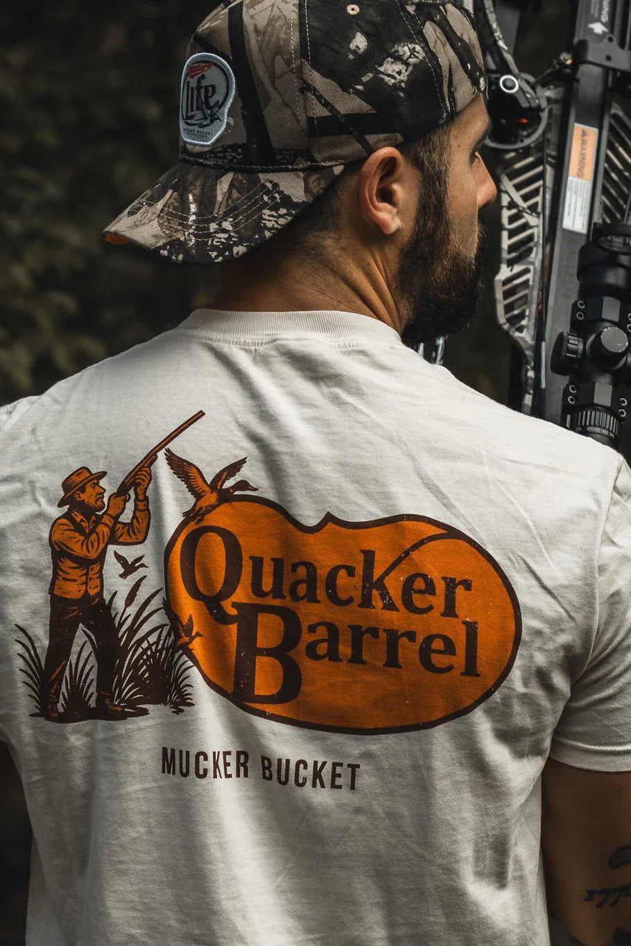 A man in a camouflage cap holds a compound bow outdoors, wearing the Mucker Bucket Quacker Barrel Premium Tee—garment-dyed cotton with a hunter, ducks, and “Quacker Barrel MUCKER BUCKET” graphic.