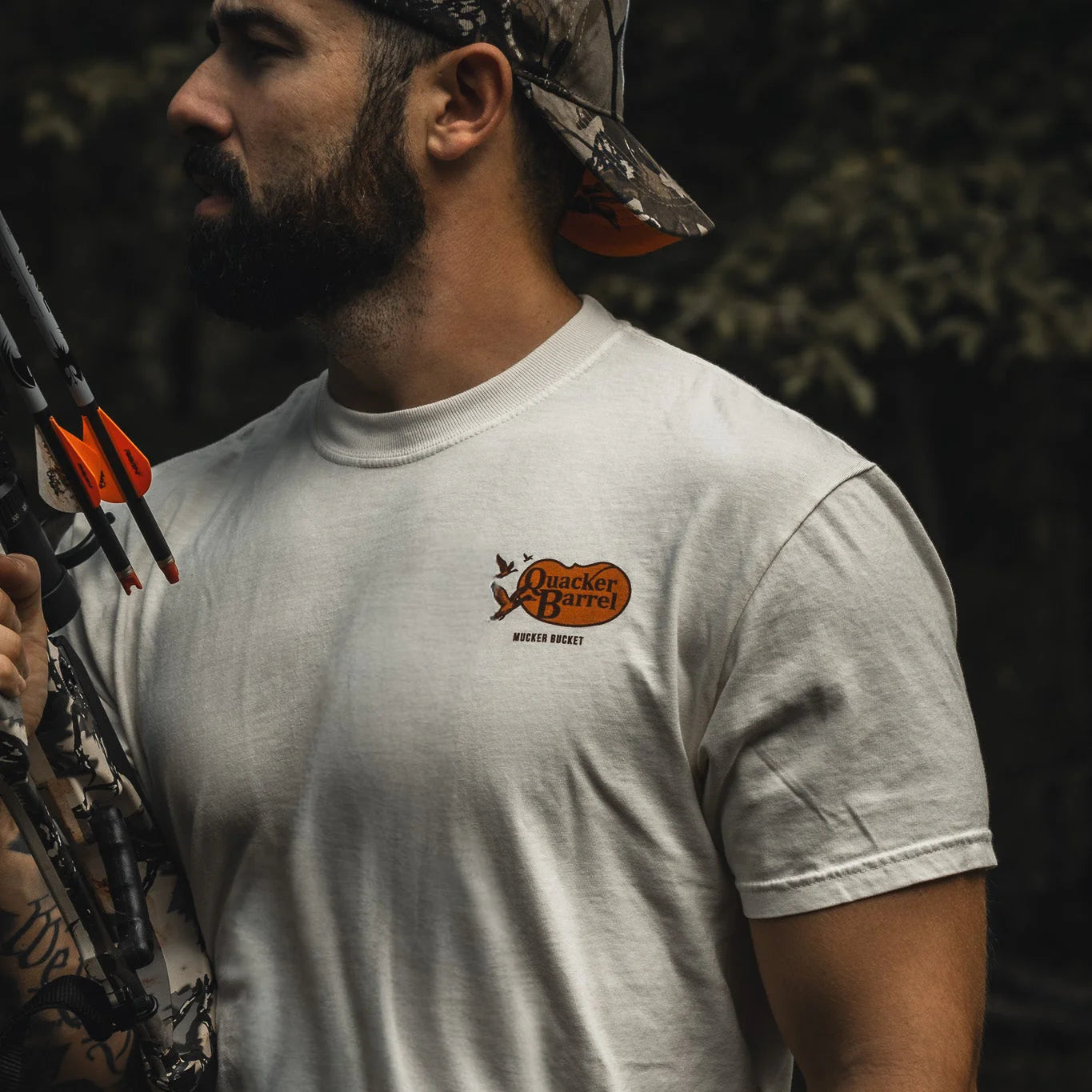 A bearded man in a Mucker Bucket Quacker Barrel Premium Tee and camo cap stands outdoors, holding a camouflage bow with orange arrows, surrounded by trees.