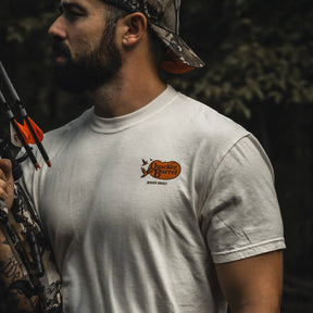 A bearded man in a Mucker Bucket Quacker Barrel Premium Tee and camo cap stands outdoors, holding a camouflage bow with orange arrows, surrounded by trees.