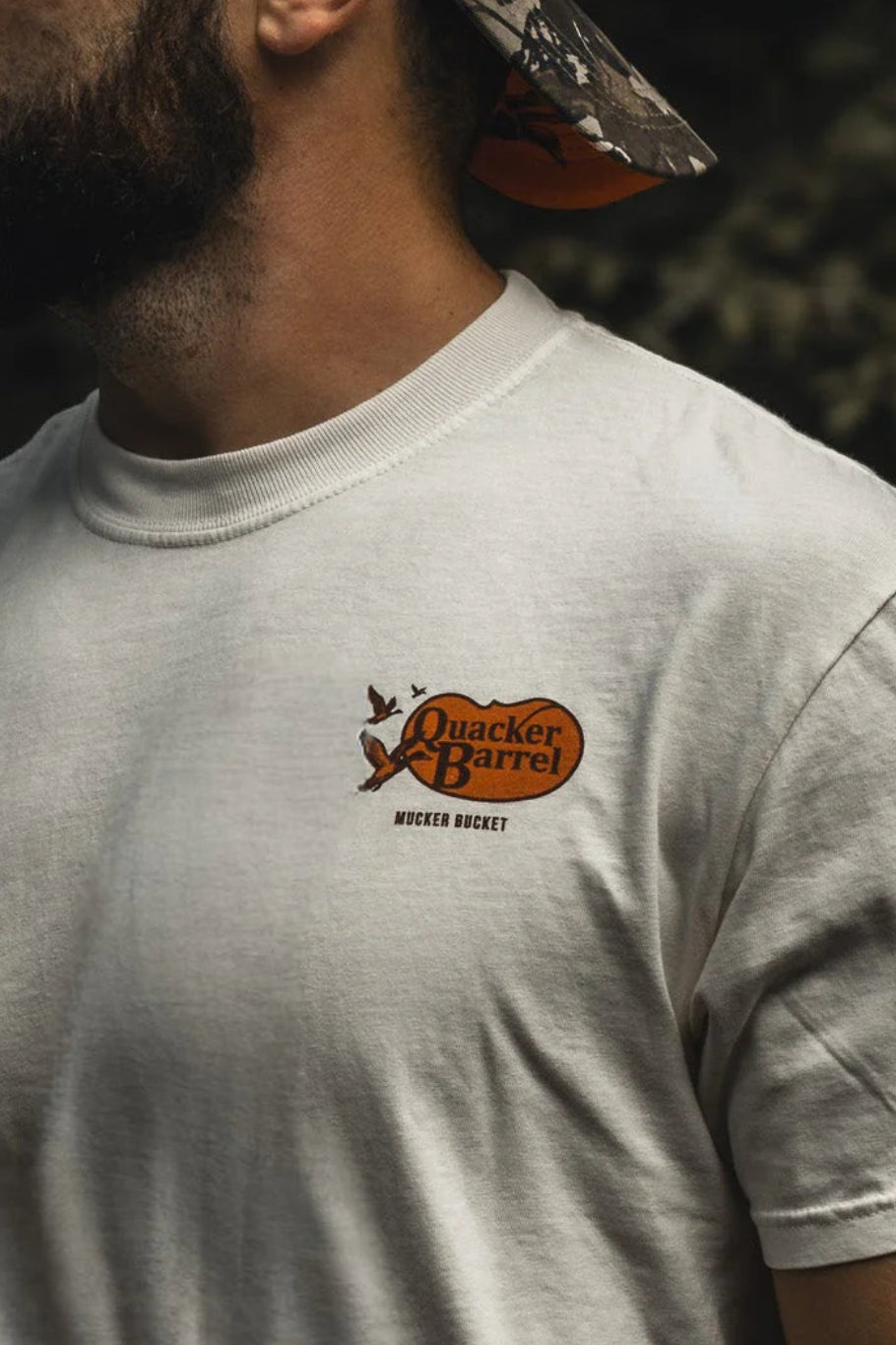 A bearded man in a Mucker Bucket Quacker Barrel Premium Tee and camo cap stands outdoors, holding a camouflage bow with orange arrows, surrounded by trees.