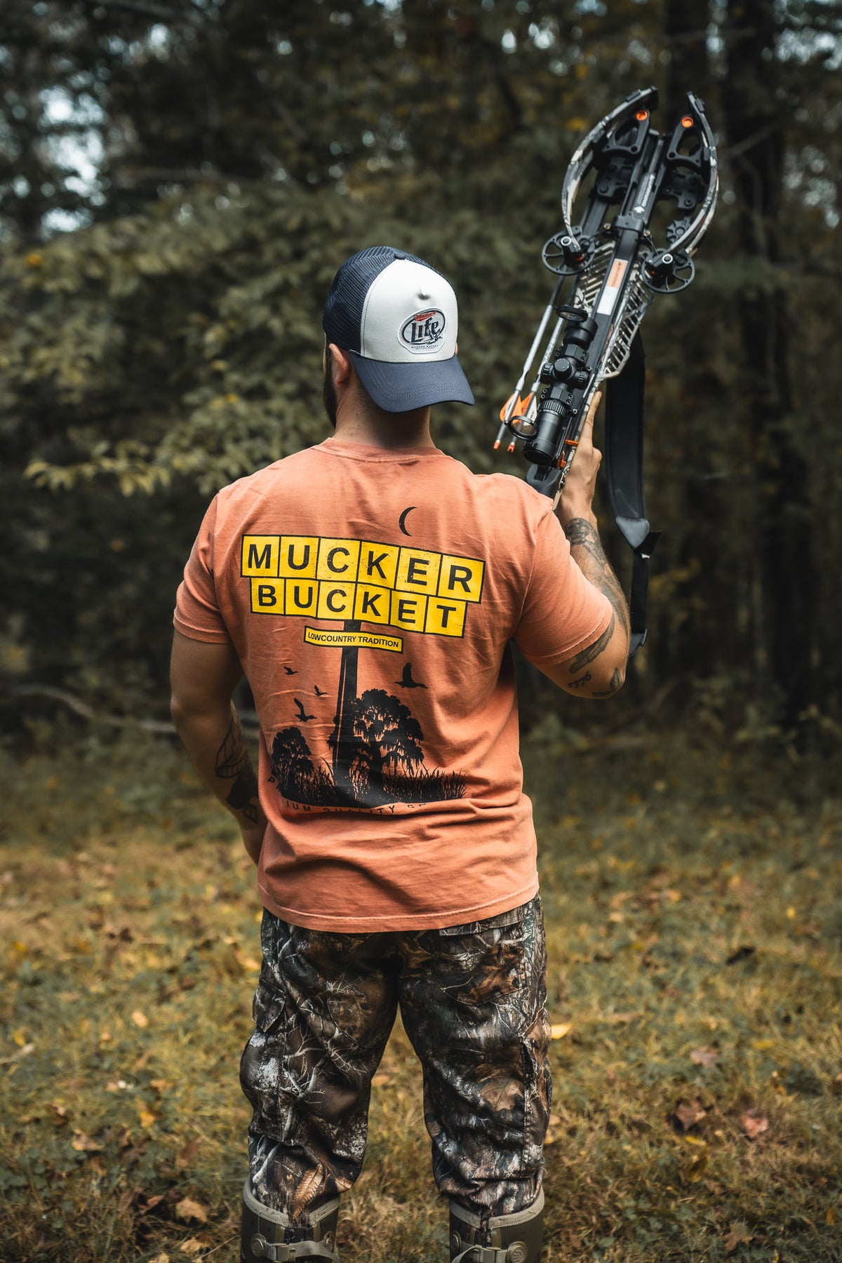 Outdoors among trees and grass, a person in camo pants, boots, and cap holds a crossbow over their shoulder. They wear the orange American Tradition Premium Tee by Mucker Bucket, featuring MUCKER BUCKET in yellow block letters.