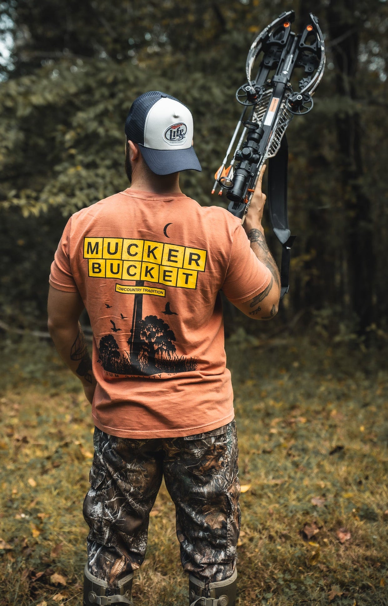 Outdoors among trees and grass, a person in camo pants, boots, and cap holds a crossbow over their shoulder. They wear the orange American Tradition Premium Tee by Mucker Bucket, featuring MUCKER BUCKET in yellow block letters.
