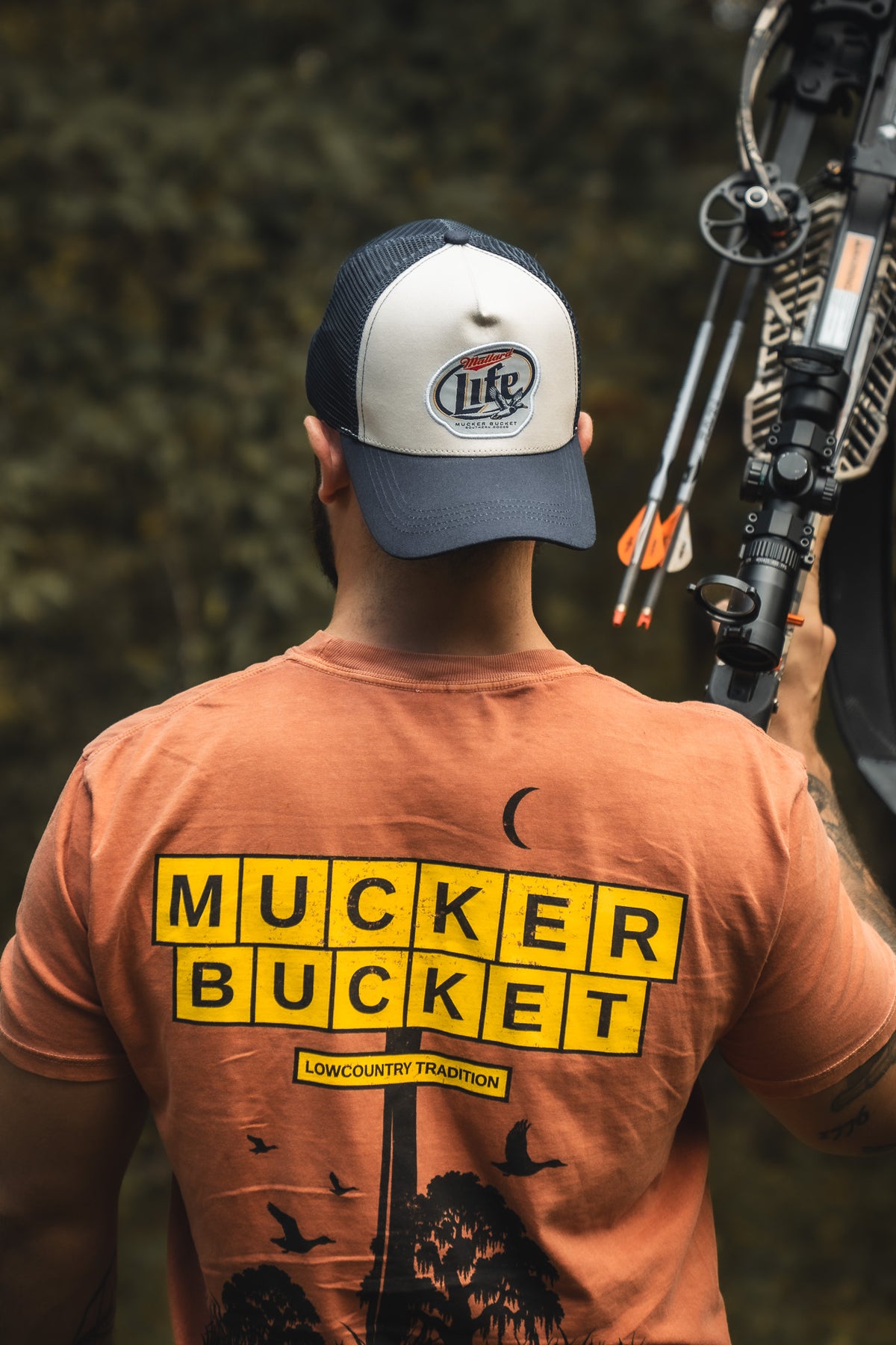 Wearing a Mucker Bucket American Tradition Premium Tee and a Miller Lite cap, a person stands outdoors with a crossbow; the photo, shot from behind, highlights the tee and gear against a softly blurred natural background.