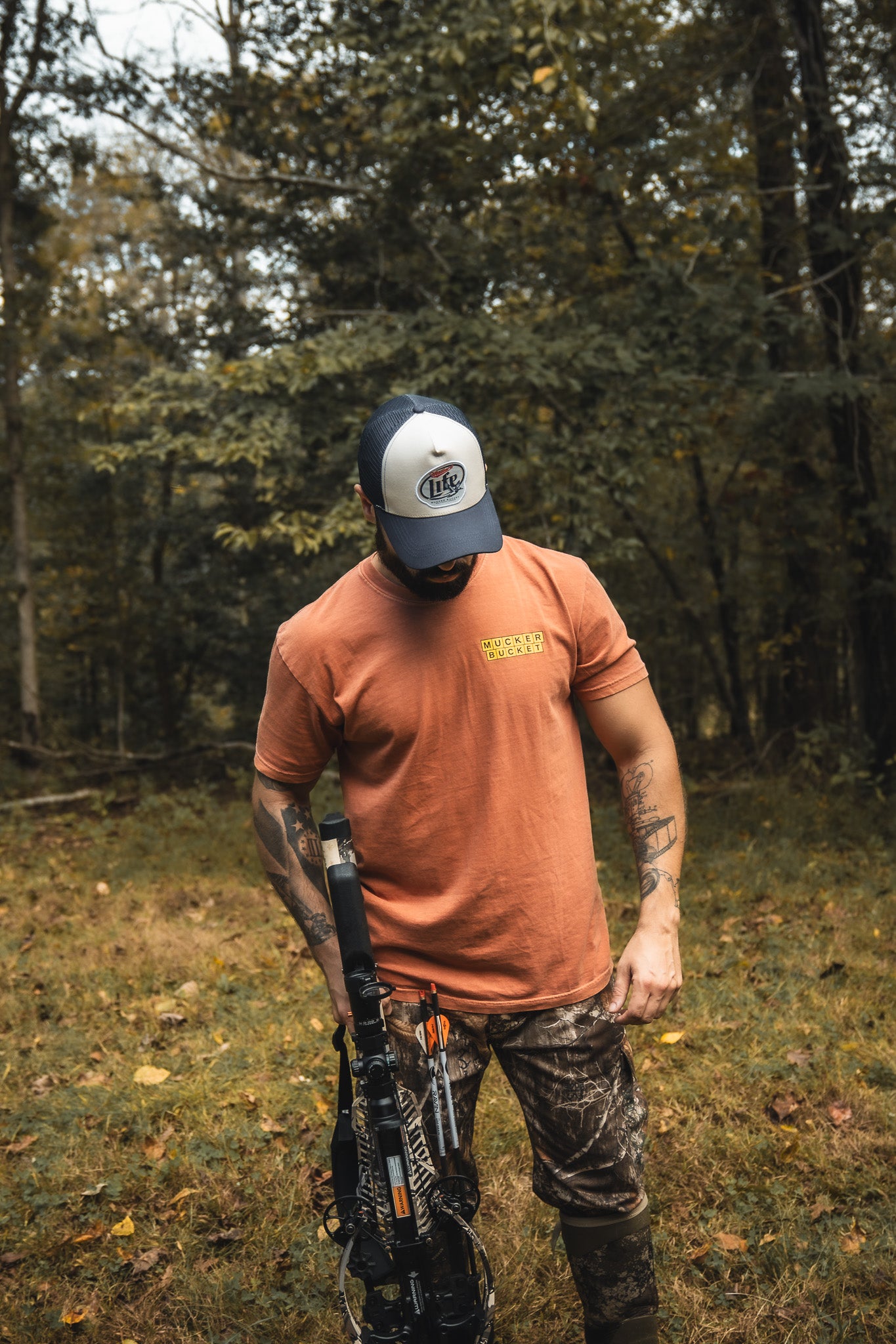 A person in a red Mucker Bucket American Tradition Premium Tee, camo pants, and a cap stands in a forest with a crossbow, looking down amid tall trees and green foliage.