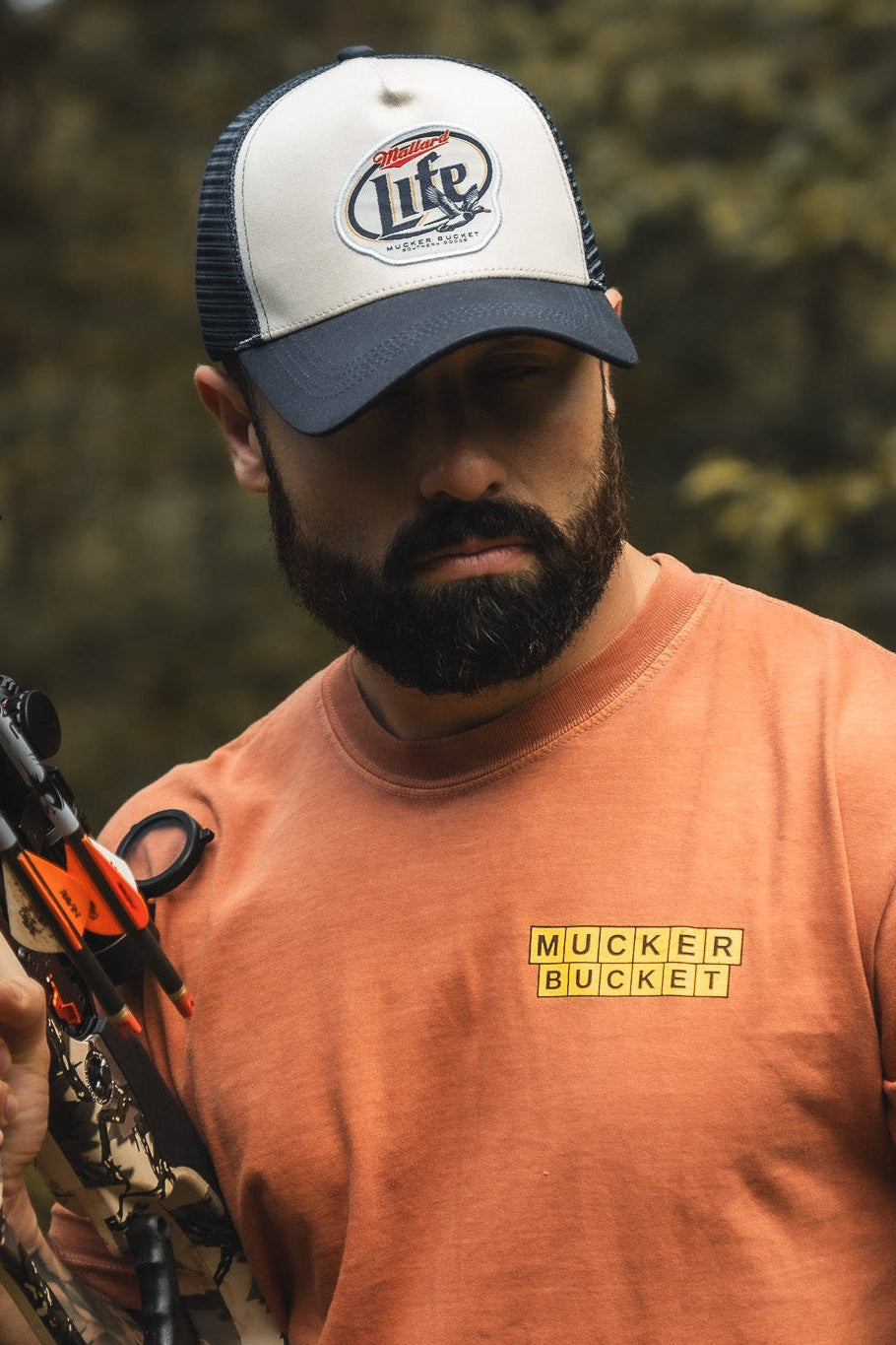 A bearded man in an orange Mucker Bucket t-shirt and a Mallard Life Trucker Bucket hat, with mesh back, holds a camouflaged crossbow outdoors amid blurred trees.
