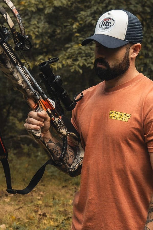 A bearded man in a Mucker Bucket "Mallard Life Trucker Bucket" hat, orange MUCKER BUCKET shirt, and holding a camouflage compound bow stands outdoors in a forested area.